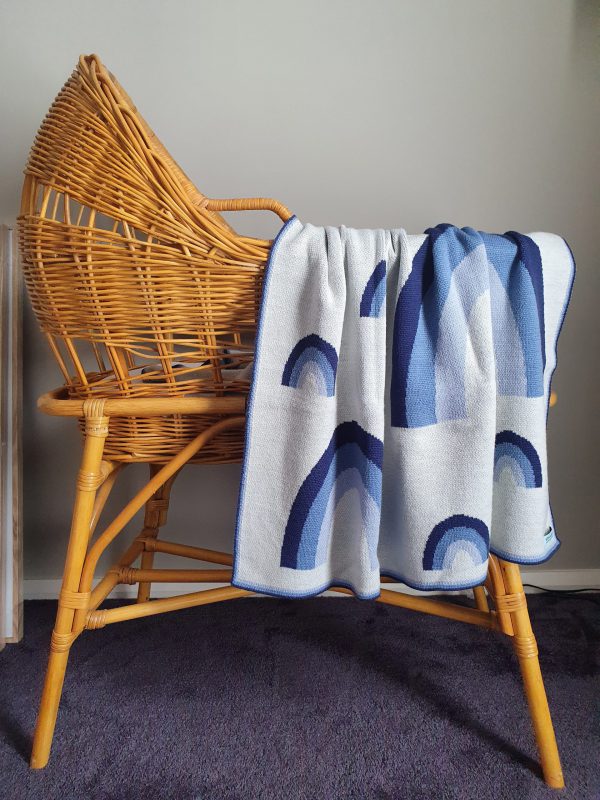Merino wool Rainbow Baby Blanket in blue shades, draped over a rattan bassinet