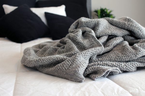Pure Merino Wool Marley Blanket in Ash (light grey), scrunched up on a bed with black and white bedding. Offers a seriously stylish addition to your bedroom.