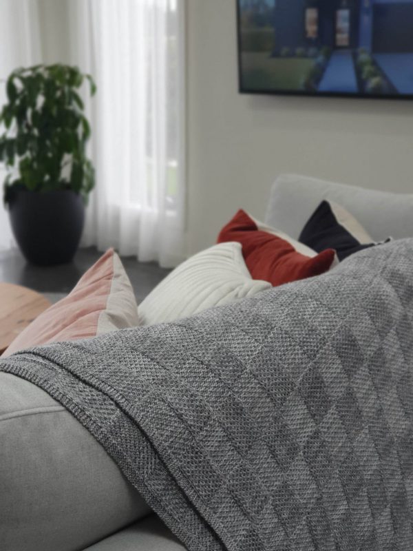 Pure Merino Wool Marley Blanket in Ash (light grey), draped over a beautifully designed couch with cushions. Viewed from the back for a stylish perspective.