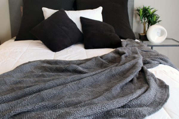 Pure Merino Wool Marley Blanket in Granite (mid grey), strewn across a double bed paired with black and white cushions. Offers a stylish bedroom setting.