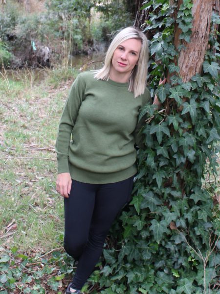 A lady leaning against a tree outside in the bush wearing a Ballarat Gold branded Australian made pure merino wool Sophie 1010 jumper in Olive green