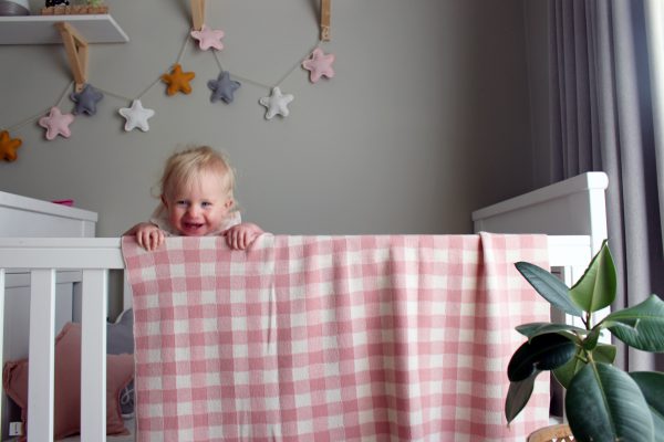 French Rose and White Gingham Blanket made from Pure Australian Merino Wool. Draped over the edge of a cot, with a matching star bunting on the wall and a happy smiling toddler girl leaning on the edge of the cot.