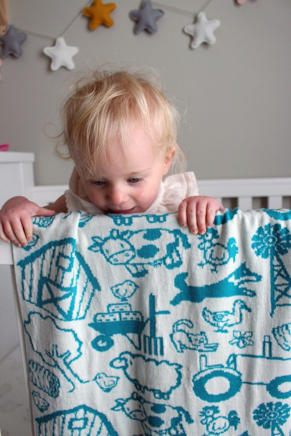 On The Farm Cot Blanket in Aqua and White, fully reversible. Close-up draped over the edge of a cot with a toddler peering over it to the floor.