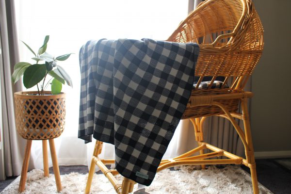Dark Navy and White Gingham Blanket made from Pure Australian Merino Wool. Nursery scene with a rattan-style bassinet in front of a window, adorned with sheer curtains and a plant in the foreground. The blanket hangs over the edge of the bassinet.