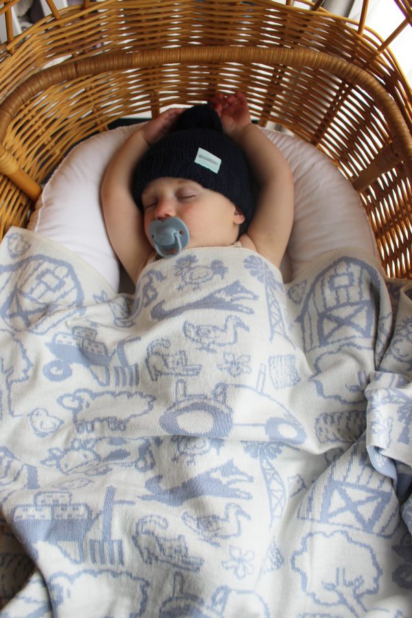On The Farm Cot Blanket in Pale Blue and White, fully reversible. Baby peacefully sleeping in a bassinet with the blanket over him, wearing our knitted wool Frankie beanie in navy.