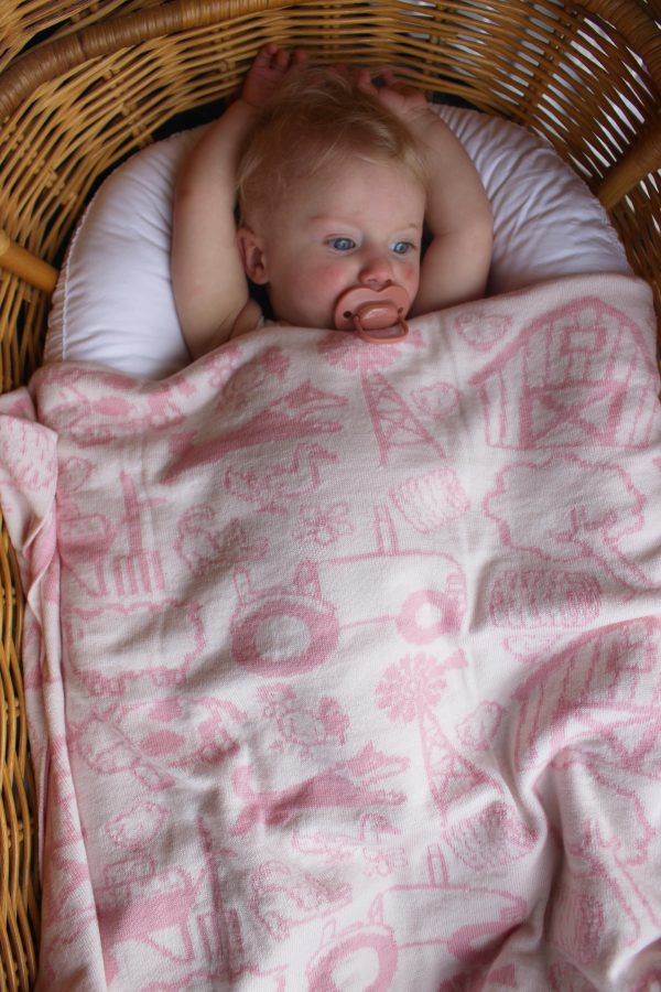On The Farm Cot Blanket - a pale pink and white reversible design. Baby restfully laying in her bassinet with the folded blanket over her.