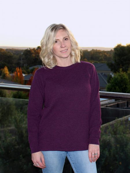 A lady standing on a balcony at sunset overlooking the trees, wearing an Australian Made, Ballarat Gold merino wool moss crew jumper in Currant (purple)