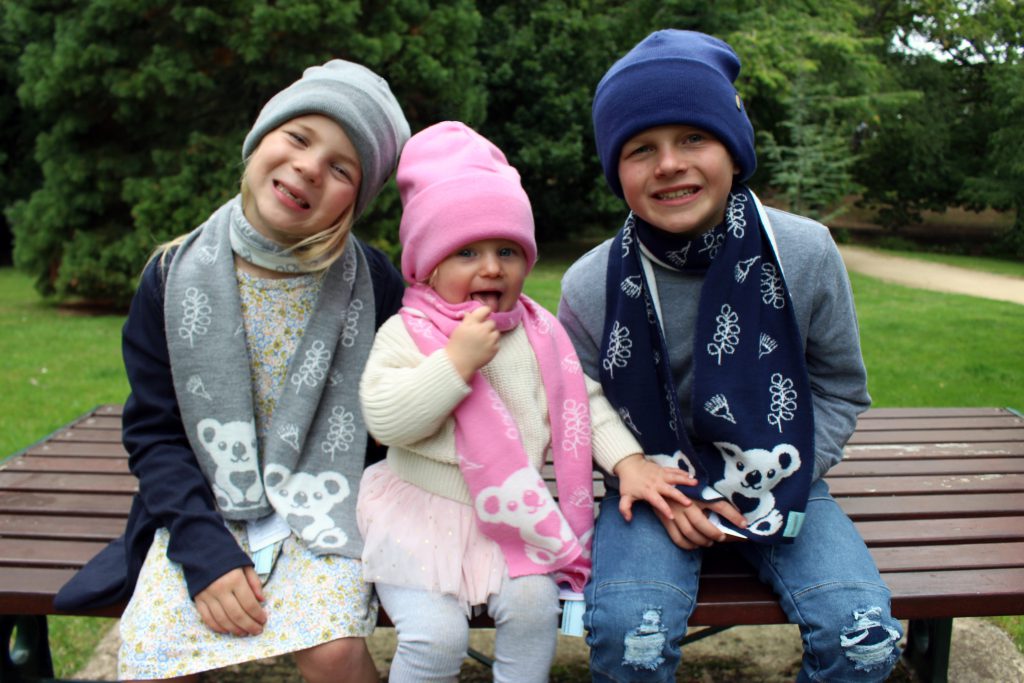 Three children sitting on a park bench wearing Branberry Australian Made, Merino wool Kids Koala Scarves and plain beanies