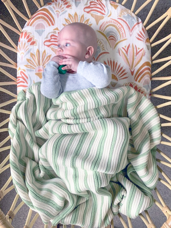 Pastel Mint Green and White Striped Garter Blanket, a gender-neutral choice, made from a luxurious blend of cotton and wool. Baby playfully moving legs in a bassinet, with the blanket messily draped over, showcasing its cosiness.