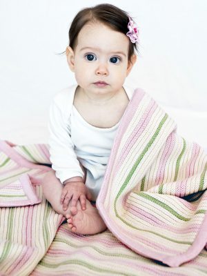 Pink, Mint, and White Garter Stripe Baby Blanket, our best-selling color combo for girls, made from a luxurious blend of cotton and wool. Toddler girl with big brown eyes sits up, snuggly draped in the soft blanket.