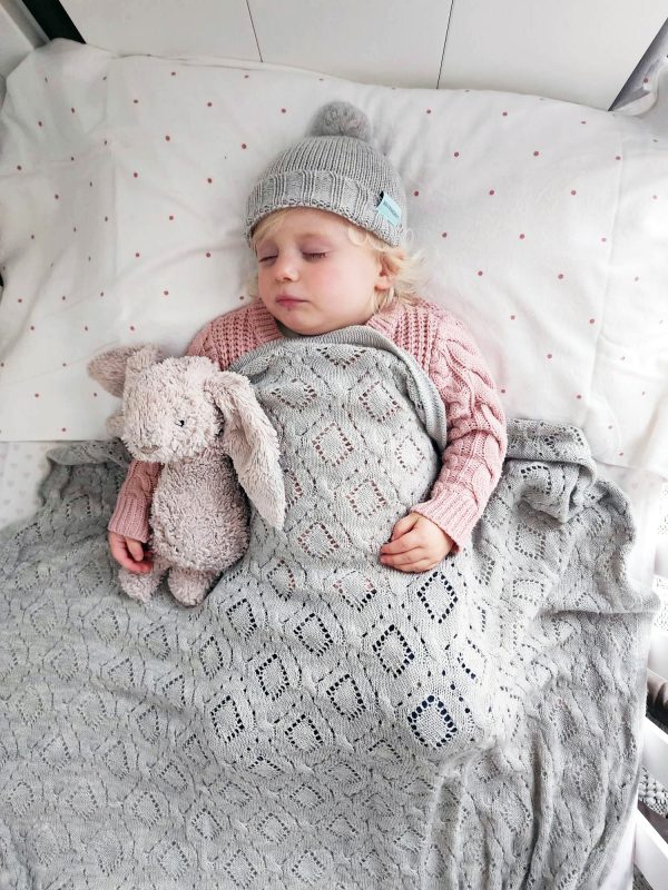 Branberry Heirloom Blanket in Smoke, a baby laying on their back, sleeping under the blanket with a matching Pure Wool Frankie Beanie and a snuggle bunny teddy
