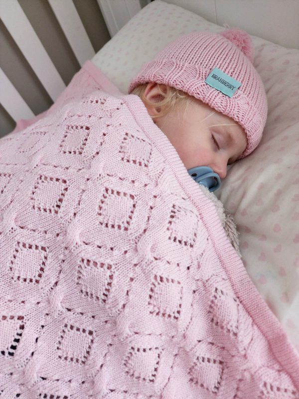 Branberry Heirloom Blanket in Pink Silk, a close-up view of a baby sleeping in a cot with the blanket, a blue dummy, and a matching Pure Wool Frankie Beanie