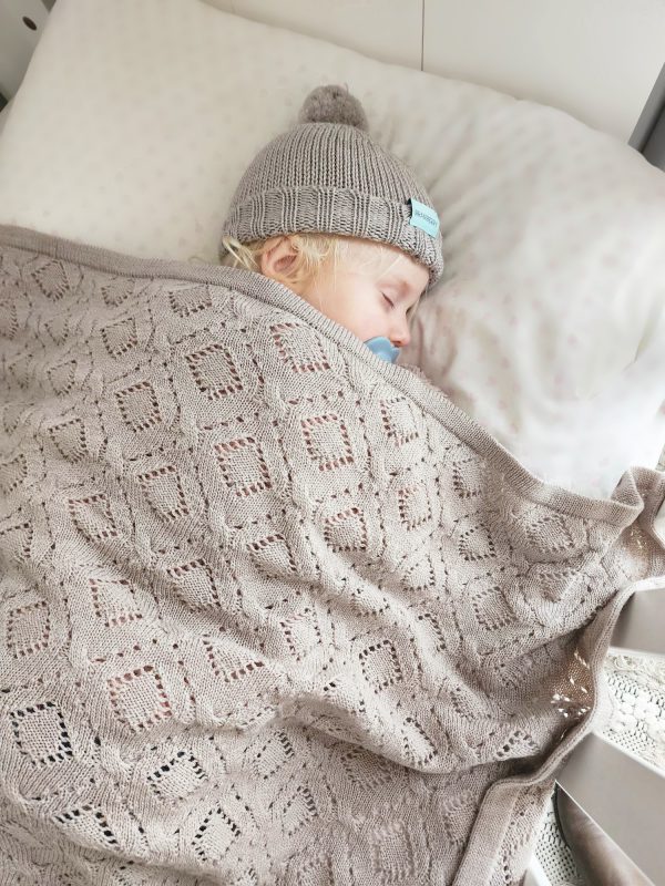 Branberry Heirloom Blanket in Cashew (beige), a baby sleeping soundly in a cot with the matching Pure Wool Frankie Beanie