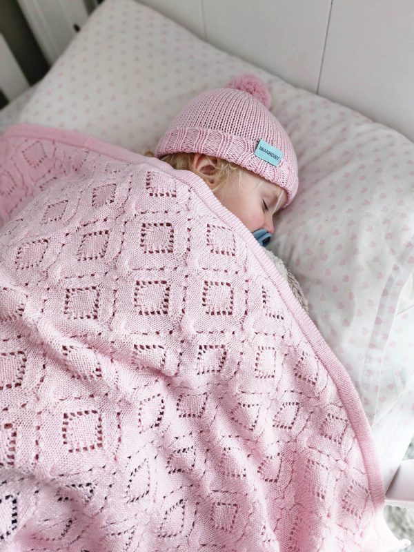 Branberry Heirloom Blanket in Pink Silk, a baby peacefully sleeping in a cot with the blanket and matching Pure Wool Frankie Beanie