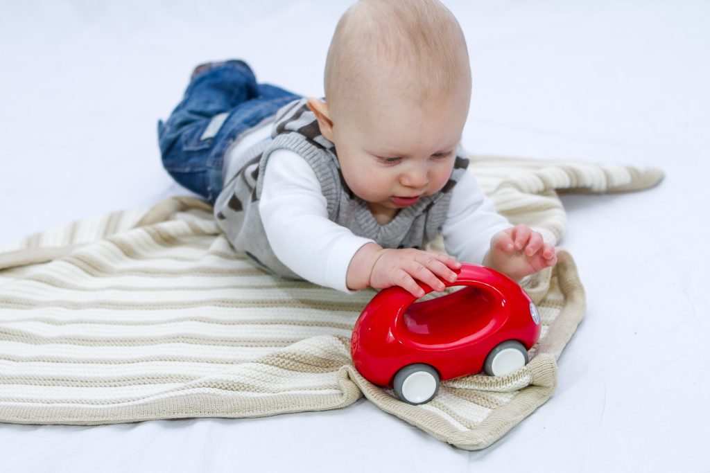 Pebble (Beige) and White Garter Stripe Baby Blanket, a serene color choice, made from a luxurious blend of cotton and wool. Baby boy plays happily with a red car toy on the floor, the blanket laid flat for his joyful exploration.