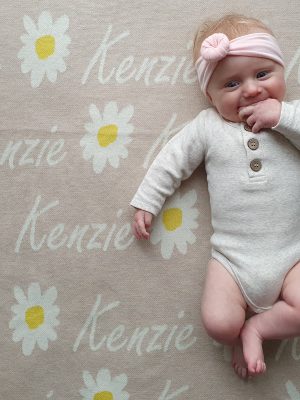Personalised Merino Wool Name Blanket in Daisy Design, a baby laying on top, happily sucking fists. Neutral Oatmeal with alternating Daisy flowers and the name 'Kenzie' in script font in white.