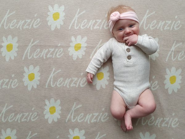 Personalised Merino Wool Name Blanket in Daisy Design, a baby laying on top, happily sucking fists. Neutral Oatmeal with alternating Daisy flowers and the name 'Kenzie' in script font in white.