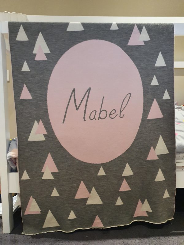 A Personalised Name Blanket with sporadic triangles with a big circle in the centre with a custom name, in a grey, pink and white colour combo, product photo. Made from Pure Australian Merino Wool and Made in Australia.
