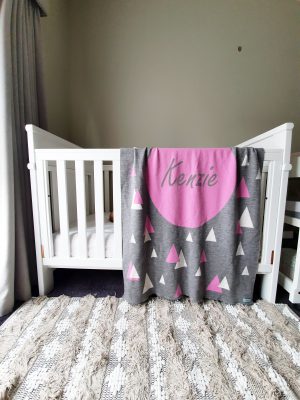 A Personalised Name Blanket with triangles with a big circle in the centre in a grey, pink and white colour combo draped over a cot. Made from Pure Australian Merino Wool and Made in Australia.