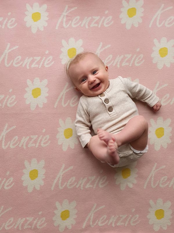 A laughing baby laying on a Personalised Name Blanket with Daisy flowers in a Pastel Pink colour. Made from Pure Australian Merino Wool and Made in Australia.