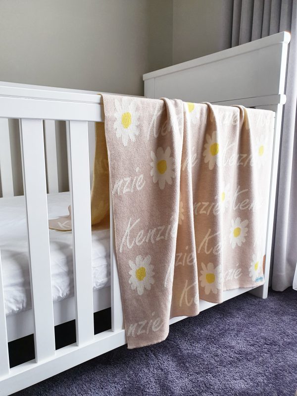 A Personalised Name Blanket with Daisy flowers in a oatmeal neutral colour draped over a cot. Made from Pure Australian Merino Wool and Made in Australia.