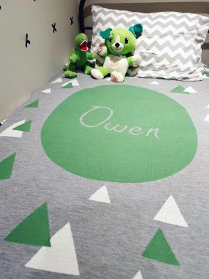 A Personalised Name Blanket with sporadic triangles with a big circle in the centre with a custom name, in a grey, green and white colour combo, placed on a bed. Made from Pure Australian Merino Wool and Made in Australia.