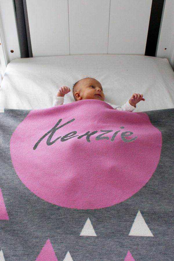 A baby tucked under her Personalised Name Blanket with sporadic triangles with a big circle in the centre with a custom name, in a grey, pink and white colour combo draped over a cot. Made from Pure Australian Merino Wool and Made in Australia.