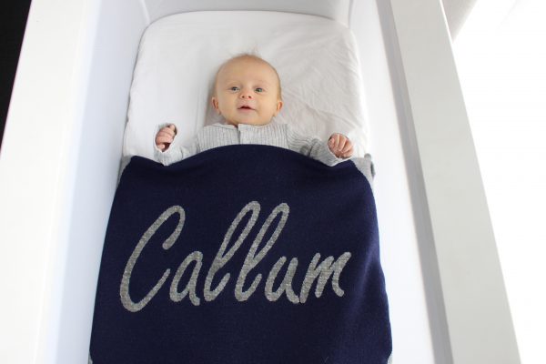 A baby tucked under a Personalised Name Blanket with sporadic triangles and a big circle in the middle with a custom name, in a Navy, Grey & White colour combo. Made from Pure Australian Merino Wool and Made in Australia.