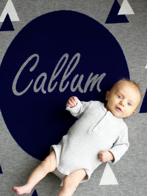 A baby laying on a Personalised Name Blanket with sporadic triangles and a big circle in the middle with a custom name, in a Navy, Grey & White colour combo. Made from Pure Australian Merino Wool and Made in Australia.