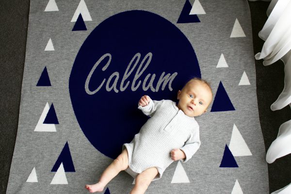 A baby laying on a Personalised Name Blanket with sporadic triangles and a big circle in the middle with a custom name, in a Navy, Grey & White colour combo. Made from Pure Australian Merino Wool and Made in Australia.