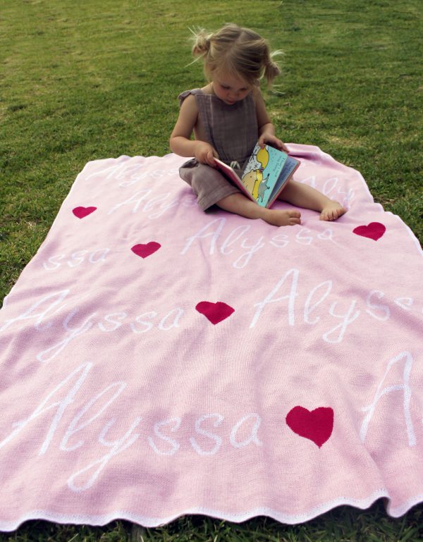 A toddler girl reading a book while sitting outside on a pure merino wool, personalised love hearts blanket in a pink, raspberry and white colour combo.