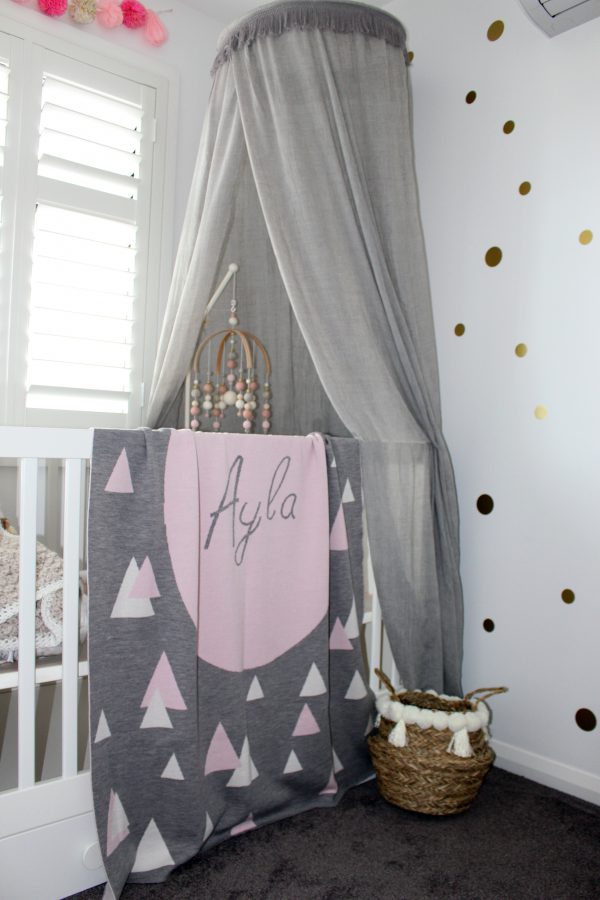 A Personalised Name Blanket with sporadic triangles with a big circle in the centre with a custom name, in a grey, pink and white colour combo draped over a cot. Made from Pure Australian Merino Wool and Made in Australia.