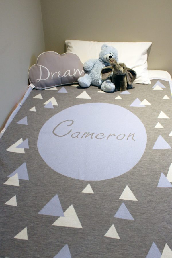 A Personalised Name Blanket with sporadic triangles with a big circle in the centre with a custom name, in a grey, blue and white colour combo, placed on a bed. Made from Pure Australian Merino Wool and Made in Australia.