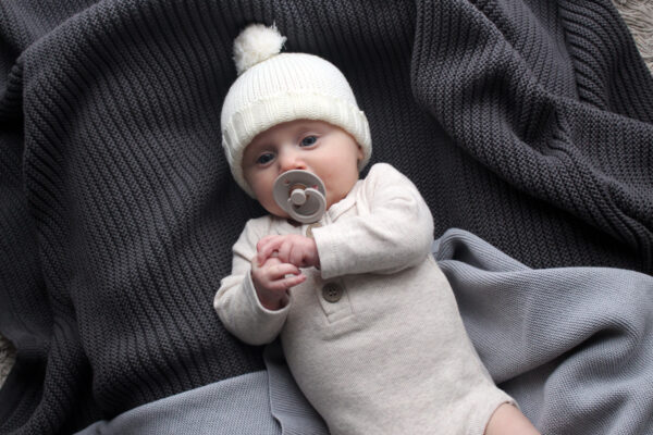 A baby laying on an Australian Made, Branberry, Plain Garter blanket in Raku Grey while wearing a Branberry Frankie beanie in Milk