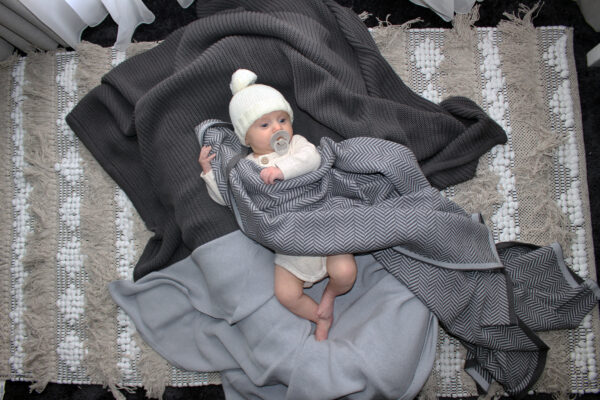 Baby laying on top of a Plain Garter Blanket in Raku grey while a Plain Garter Blanket in Grey is draped over the baby. The baby is wearing a Branberry Frankie beanie in Milk