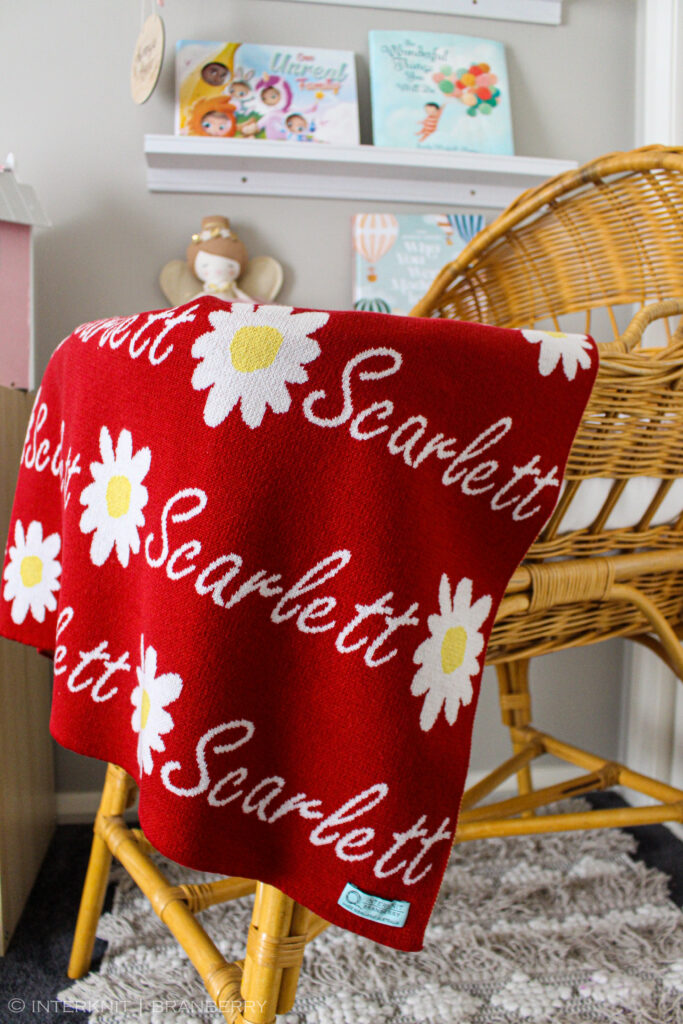 Australian Made, Merino Woollen Personalised Daisy Knitted Baby Blanket in Sangria Red, draped over the edge of a bassinet in a nursery. Features alternating an Daisy flower and a personalised name knitted in a script font.