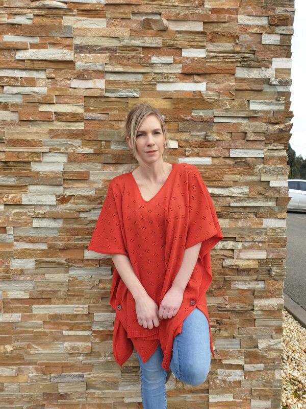 A woman leaning against a stone wall wearing the Harlow Poncho in coral, exuding relaxed elegance.