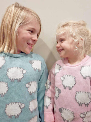 Two children standing indoors wearing a merino wool jumper with sheep patterns, smiling happily at each other.