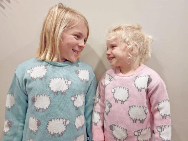 Two children standing indoors wearing a merino wool jumper with sheep patterns, smiling happily at each other.