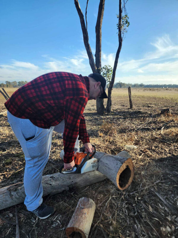 Man chopping wood with a chainsaw in a red and black Woolly Camper shirt jumper in a dry, desert-like paddock.