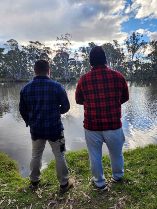 Two men wearing blue/black and red/black Woolly Camper shirt jumpers standing by a lake at dusk.