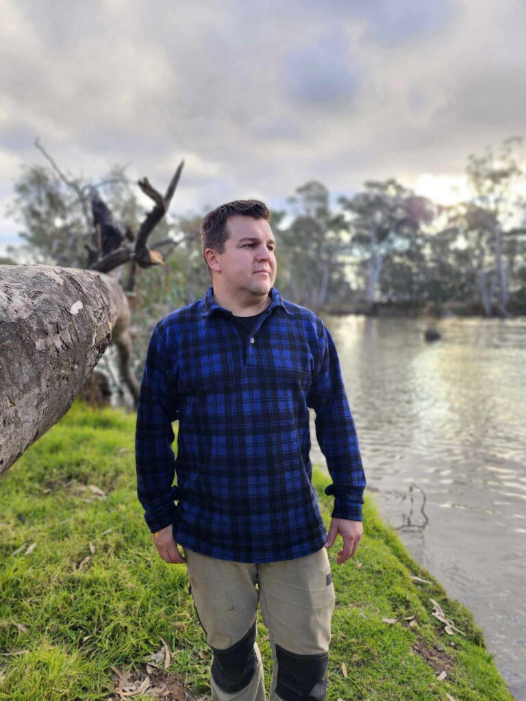 Man wearing blue and black check Woolly Camper shirt jumper by a lake at dusk, surrounded by greenery.