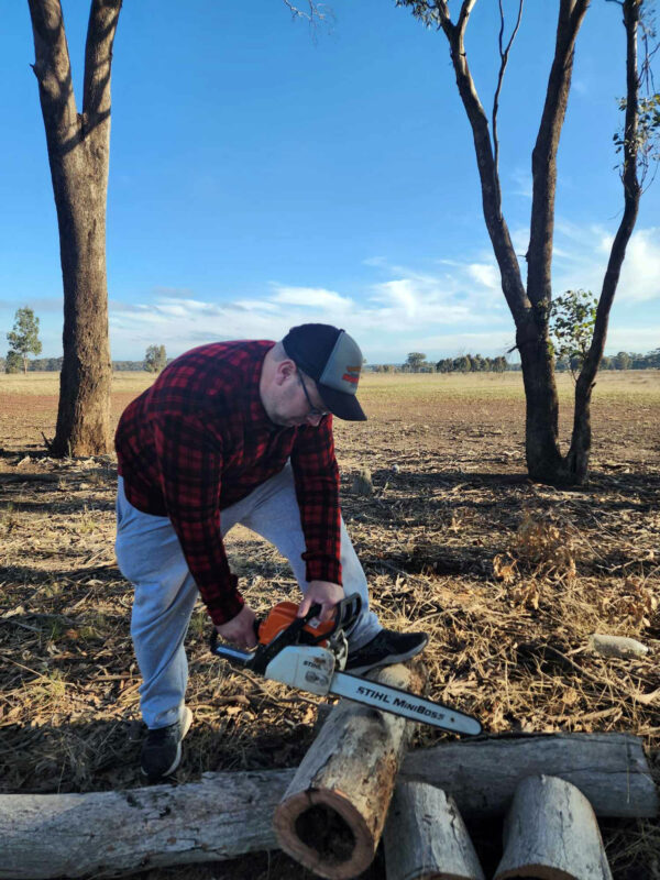 Man wearing red and black Woolly Camper shirt jumper, chopping wood with a chainsaw in a dry paddock under a bright sky.